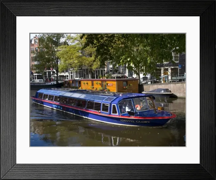 Framed Print : A glass topped tour boat travels down the Amstel river lined with trees and houseboats Framed Print : A glass topped tour boat travels down the Amstel river lined with trees and houseboats