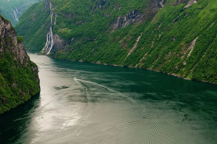 Framed Print : A boat's wake ripples Geirangerfjord as it winds through forest covered sheer cliffs and mountains, Seven Sisters waterfalls is in the distance. Geirangerfjord, Norway