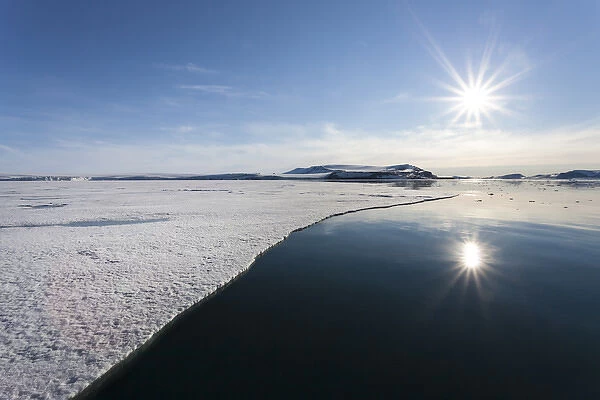 Europe, Svalbard. Sunburst reflected in still water at edge of shorefast ice. Credit as