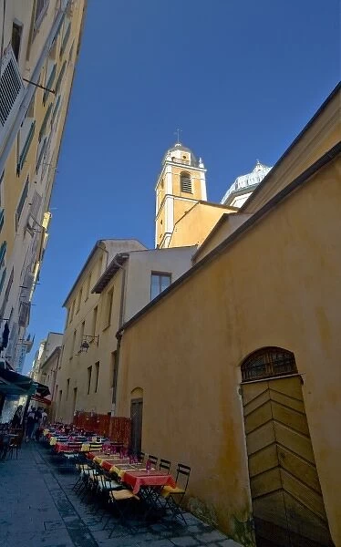 France, Corsica. Church steeple rises above narrow street with tables set up outside