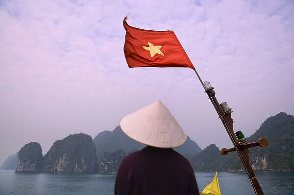 Girl with conical hat on a junk boat with national flag and karst islands in Halong Bay