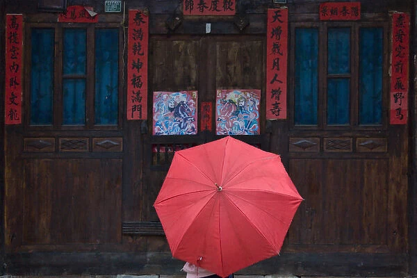 Girl with red umbrella watching traditional wood door decorated by poster of guarding god