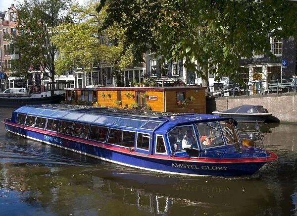A glass topped tour boat travels down the Amstel river lined with trees and houseboats