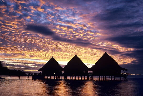 A spectacular sunset silhouettes a group of thatched-roof overwater bungalows in