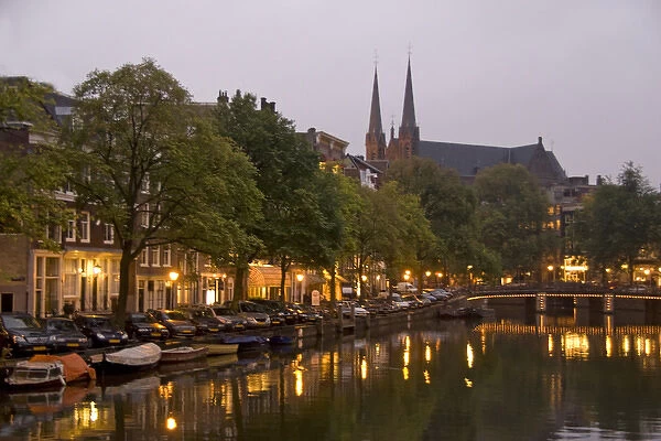 Street lights reflect onto the canal at dusk with a lit bridge and De Krijtberg in