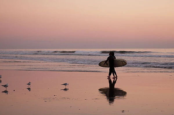 Surfers cast a shadow on wet sand as they walk on Huntington Beach at sunset. California, USA
