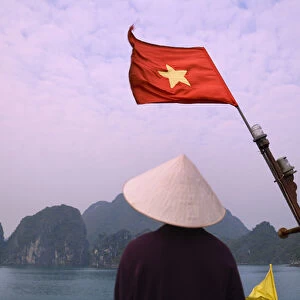 Girl with conical hat on a junk boat with national flag and karst islands in Halong Bay