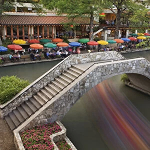 Outdoor cafe along River Walk and boat in motion passing under bridge over San Antonio River