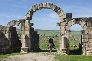 Ruins Prints: Africa, Morocco. A man on a donkey passes under stone columns and arches at the roman