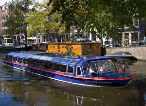 A glass topped tour boat travels down the Amstel river lined with trees and houseboats A glass topped tour boat travels down the Amstel river lined with trees and houseboats