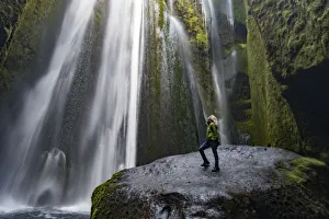 Prints of Iceland, Seljalandsfoss, woman standing on large rock in front of streaming falls in green slot canyon. (MR) Images Dated 2019 January: Iceland, Seljalandsfoss, woman standing on large rock in front of streaming falls