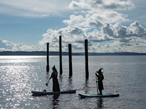 Prints of Port Townsend, Washington State. Silhouettes of two women dressed as witches on paddleboards Images Dated 2024 October: Port Townsend, Washington State