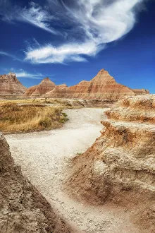 Badland Prints: Sandy path leads through the striated peaks to the higher peaks in the background stark against a blue sky