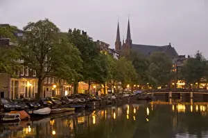 Marilyn Parver Prints: Street lights reflect onto the canal at dusk with a lit bridge and De Krijtberg in