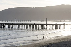Relaxed Prints: US, CA, Avila Beach. Silhouetted beach walkers approach pier end of day