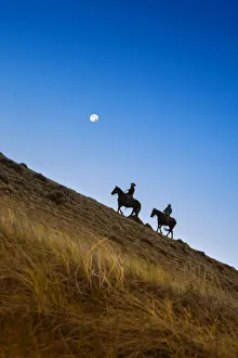Golden Prints: Two wranglers riding horses up a hill with full moon in backround at blue hour